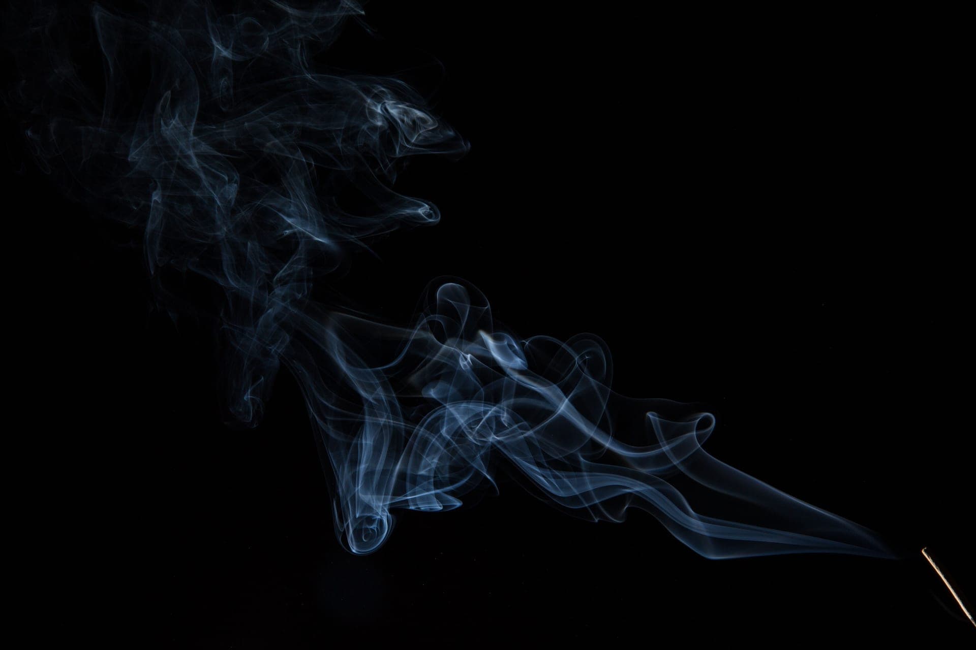 A thin ribbon of smoke from an incense stick bending sideways next to a window frame